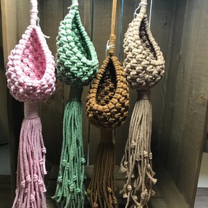 May include: Four macrame plant hangers in pink, green, brown, and beige. Each features a knotted basket design with a long tassel. The hangers are suspended against a wooden background, showcasing a bohemian style.
