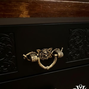 Könnte beinhalten: Nahaufnahme einer schwarzen Schublade mit einem dekorativen Messinggriff. Der Griff zeigt einen Totenkopf in einem Rosendesign. Die Schublade hat verzierte Schnitzereien und den Text "Dusk+Dawn Votum".