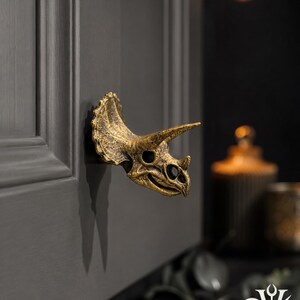 May include: A gold-toned cabinet knob shaped like a triceratops skull with a textured, weathered finish. The knob is mounted on a dark gray cabinet door. The text DUSK+DAWN VOTUM is visible in the upper left corner of the image.