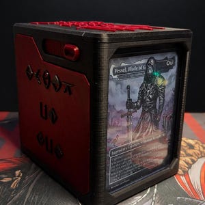 May include: A black and red rectangular deck box featuring a clear front display window holding a trading card titled Vessel, Blade of G. The side panel displays raised black symbols, and the top lid has red lettering in a stylized font.