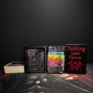 May include: A black deck box with a card featuring a knight, a holographic trading card, and a black notebook with the red embroidered text "Nothing Lasts Forever" and a red symbol. A silver chain necklace with a small shell pendant rests in front.