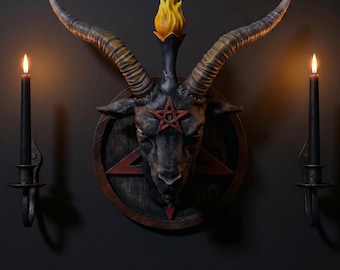 May include: A Baphomet plaque featuring a dark goat head with large curved horns and a yellow flame at the top. A red pentagram is centered on the forehead and on the circular wooden backing. Two black candle holders with lit candles flank the plaque.
