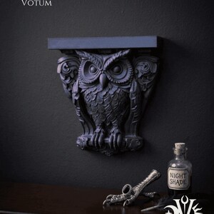 May include: A dark gray owl-shaped wall shelf with intricate details, mounted on a dark wall. The shelf has a flat top and is flanked by decorative carvings. The image also includes a small bottle labeled "NIGHT SHADE" and a decorative bird claw.