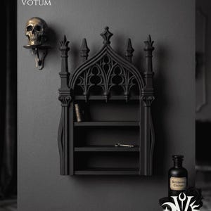 May include: A black gothic-style wall shelf with three shelves, a decorative top, and a key. A golden skull on a wall-mounted shelf is to the left. A black bottle and a decorative stand with a logo are on the right. The text "Dusk + Dawn Votum" is at the top.