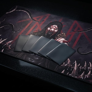 May include: A large rectangular desk mat featuring a dark illustration of a hooded figure with red facial markings, surrounded by tentacles and sharp teeth, with five plain black rectangular cards layered in a fan shape on top of the mat.