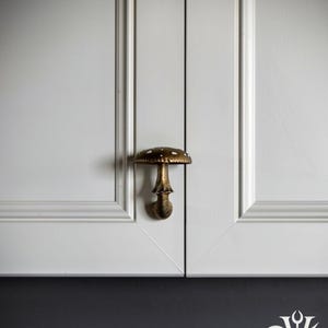 May include: A brass-toned mushroom-shaped cabinet knob with white spots on the cap, mounted on a white cabinet door. The text DUSK+DAWN VOTUM appears in the top left corner, and a stylized logo with a moon and sun is in the bottom right.