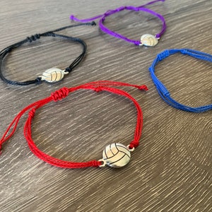 May include: Four colorful string bracelets with a silver volleyball charm. The bracelets are red, black, purple, and blue.