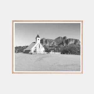May include: A black and white photograph of a small white church with a steeple and a cross on top. The church is in front of a mountain range.