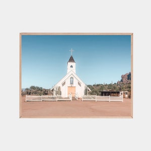 May include: A white church with a wooden door and a cross on the steeple stands in a desert landscape. The church is surrounded by a white picket fence and there are mountains in the background.