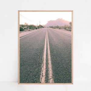 May include: A long, straight road with a white double line down the center, leading towards a mountain range in the distance. The road is surrounded by desert vegetation and the sky is a soft pink color.