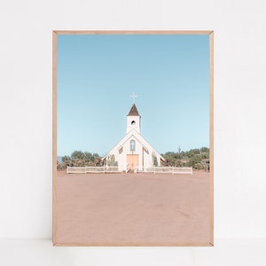 May include: A white church with a wooden cross on the roof, surrounded by desert vegetation and a white picket fence. The church is set against a bright blue sky.