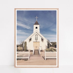 May include: A white church with a wooden cross on the roof, surrounded by a white picket fence and cacti. The church is set against a blue sky with white clouds.
