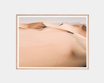 May include: A photograph of a desert landscape with rolling sand dunes. The sand is a light beige color and there are footprints in the sand.