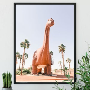 May include: A large, brown dinosaur statue stands in a desert landscape with palm trees. The statue is made of concrete and has a long neck and tail. The dinosaur is facing forward and its mouth is slightly open.