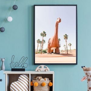 May include: A framed print of a large, brown dinosaur statue in a desert setting with palm trees. The dinosaur is standing on a dirt path with a blue sky in the background.