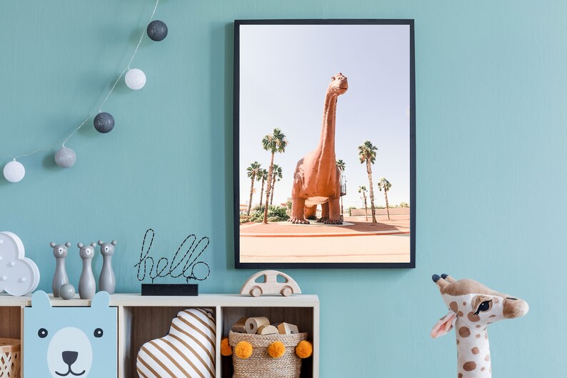 May include: A framed print of a large, brown dinosaur statue in a desert setting with palm trees. The dinosaur is standing on a dirt path with a blue sky in the background.