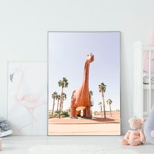 May include: A framed print of a large, brown dinosaur standing in a desert landscape with palm trees. The dinosaur is facing forward with its head tilted up. The sky is blue and the sand is a light brown.