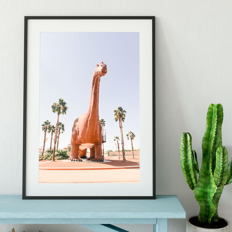 May include: A framed photograph of a large, brown dinosaur statue in a desert setting. The dinosaur is standing tall with its head tilted up, and there are palm trees in the background.