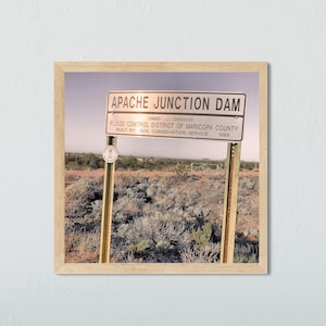 May include: A sign for the Apache Junction Dam, which was built in 1988 by the Soil Conservation Service. The sign is located in a desert landscape with brown dirt and sparse vegetation.