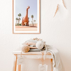 May include: A framed photograph of a large, pink dinosaur statue in front of a row of palm trees. The photo is hanging on a white wall above a wooden stool with a stack of knitted blankets and a tag.