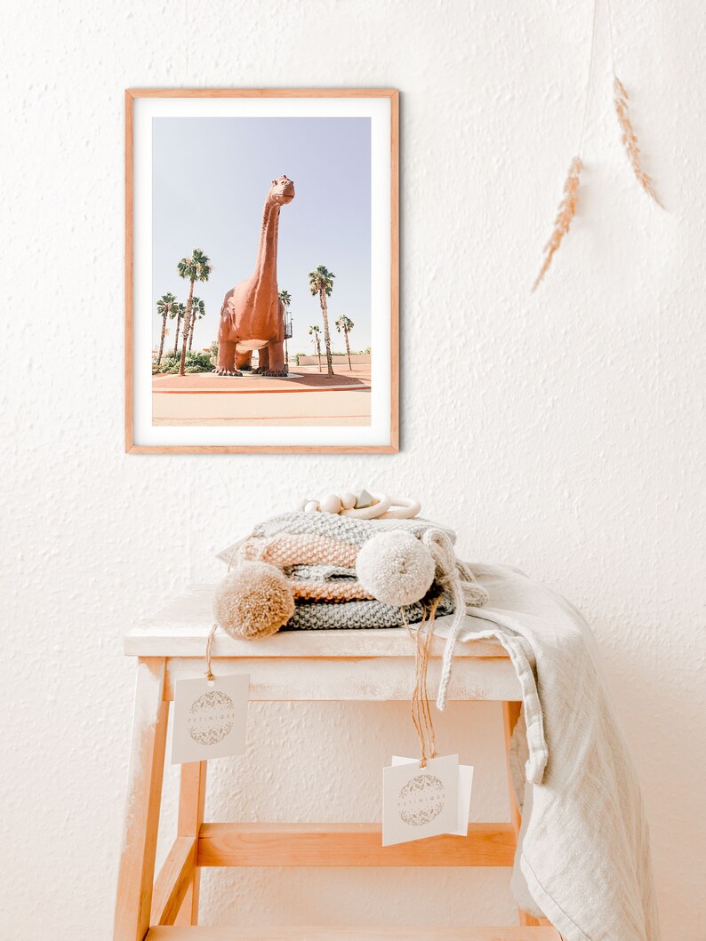 May include: A framed photograph of a large, pink dinosaur statue in front of a row of palm trees. The photo is hanging on a white wall above a wooden stool with a stack of knitted blankets and a tag.