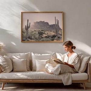 May include: A framed landscape art print of a desert scene with cacti and mountains hangs above a light-colored wooden sofa with white cushions. A person is seated on the sofa, reading a book. The room has a neutral color palette.