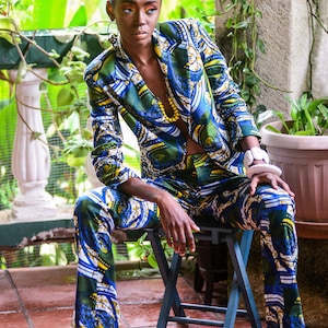 Puede incluir: Un traje estampado en azul, verde y amarillo, compuesto por una americana y un pantal&oacute;n. El traje se lleva con un collar de cuentas amarillas y una pulsera blanca. La persona est&aacute; sentada en un peque&ntilde;o taburete azul, con un fondo verde exuberante.
