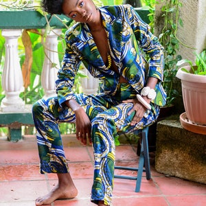Puede incluir: Un traje estampado en tonos azules, verdes y amarillos. El traje consta de una americana y un pantal&oacute;n a juego. La persona est&aacute; sentada en un peque&ntilde;o taburete azul en un patio de baldosas rojas, rodeado de vegetaci&oacute;n.