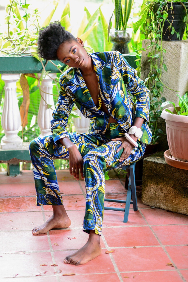 Puede incluir: Un traje estampado en tonos azules, verdes y amarillos. El traje consta de una americana y un pantal&oacute;n a juego. La persona est&aacute; sentada en un peque&ntilde;o taburete azul en un patio de baldosas rojas, rodeado de vegetaci&oacute;n.