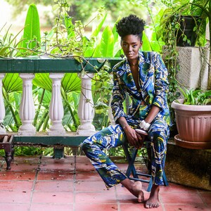 Puede incluir: Una persona con un traje estampado en azul, verde y amarillo, sentada en un taburete peque&ntilde;o en un patio embaldosado. El traje tiene un dise&ntilde;o abstracto &uacute;nico. El fondo incluye exuberante vegetaci&oacute;n y una balaustrada blanca.