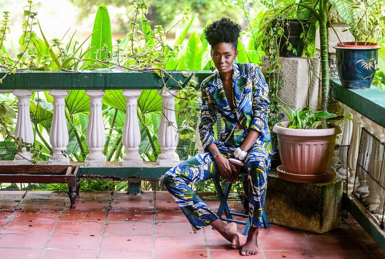Puede incluir: Una persona con un traje estampado en azul, verde y amarillo, sentada en un taburete peque&ntilde;o en un patio embaldosado. El traje tiene un dise&ntilde;o abstracto &uacute;nico. El fondo incluye exuberante vegetaci&oacute;n y una balaustrada blanca.