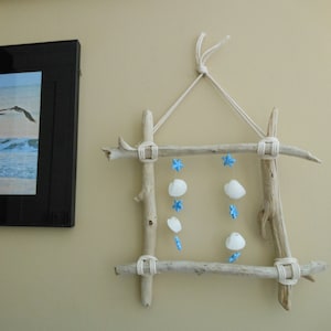 May include: A driftwood frame with white seashells and blue star-shaped charms hanging from it. The frame is hung on a light beige wall.