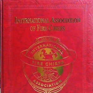 May include: A red book with gold lettering on the cover. The title is "International Association of Fire Chiefs" and "Commemorative History". The cover also features a gold emblem with a globe and the words "International Fire Chiefs Association".