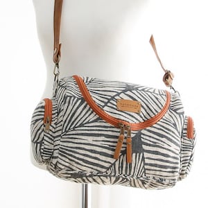 May include: A black and white patterned canvas shoulder bag with a brown leather strap and zipper closures. The bag has a small pocket on the front and the text "handmade" on a brown leather tag.