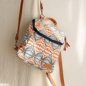 May include: A patterned backpack with a brown leather handle and straps. The backpack features a repeating leaf-like design in orange, blue, and white. The bag has a zipper closure and a small tag that says "handmade".