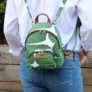 May include: A small backpack featuring a green and white abstract pattern with a textured fabric finish. It includes a brown leather top handle, dark brown zippers, and adjustable green shoulder straps. A small front pocket displays a rectangular tag with the text HAND MADE.