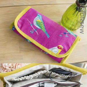May include: A magenta fabric organizer with a yellow trim and a bird print. The organizer is open, revealing multiple compartments with a zip closure, holding a phone, charger, and cable. The organizer is on a wooden surface.