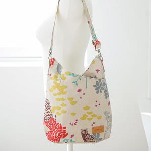May include: A beige hobo bag with a colorful floral and owl print featuring red, yellow, and gray patterns. The bag has a single shoulder strap, a brown leather-like tag labeled handmade, and the text ithinksew printed in the upper right corner.