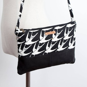 May include: Black and white crossbody bag with a repeating pattern of white birds on a black background. The bag has a black leather bottom and a brown leather tag that says "HANDMADE".
