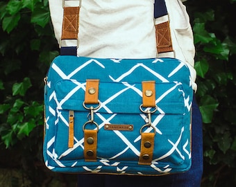 Può includere: Una borsa a tracolla blu e bianca con motivo, con dettagli in pelle marrone e una tasca con cerniera. La borsa ha due anelli metallici e il testo "HANDMADE" stampato sul davanti.