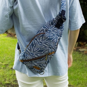 Op de afbeelding: Een marineblauwe sling bag met een wit stippenpatroon. De tas heeft een bruine rits en afwerking, en een zwarte verstelbare riem. De tas wordt over het lichaam gedragen, met de riem over de schouder.