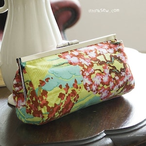 Puede incluir: Una jarra de cerámica blanca con un diseño floral se encuentra sobre una mesa de madera. La jarra está llena de rosas blancas. Un embrague de tela colorido con un patrón floral está delante de la jarra.