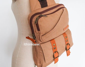 May include: A tan canvas crossbody bag with multiple pockets and a brown strap. The bag has two buckles with orange leather straps and metal accents. The bag has a zipper closure and the text "ithinksew" is visible on the side.