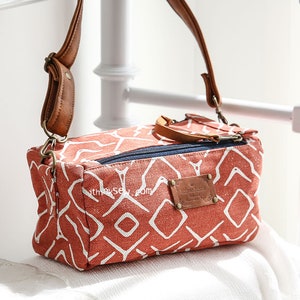 Puede incluir: Un pequeño bolso bandolera rectangular con una correa de cuero marrón y un cierre de cremallera. El bolso está hecho de lona y tiene un patrón geométrico en tonos naranja y blanco. El bolso tiene un parche de cuero con el texto "Vintage Style" en él.