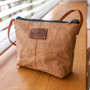 以下が含まれることがあります： しわくちゃの紙のような素材で作られた茶色のクロスボディバッグ。革のストラップとジッパーの留め具が付いています。バッグには、「The Journey」という文字が書かれた茶色の革のパッチが付いています。