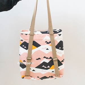 Puede incluir: Una bolsa de mano rosa y blanca con un patrón geométrico de montaña en negro, blanco y amarillo. La bolsa tiene correas marrones y un asa de cuero marrón.
