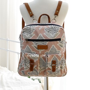 Könnte beinhalten: Ein rosa-weißer Rucksack mit floralem Muster, braunen Lederriemen und Schnallen. Der Rucksack hat zwei Fronttaschen mit Schnallen und einen Reißverschluss oben. Der Text "i think sew.com" ist auf der Vorderseite der Tasche aufgedruckt.