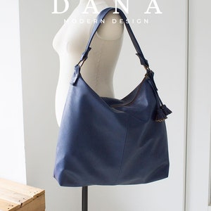 Könnte beinhalten: Eine dunkelblaue Ledertasche mit einem langen Trageriemen und einem Quastenanhänger am Reißverschluss. Die Tasche befindet sich auf einem weißen Mannequin. Der Text "DANA MODERN DESIGN" befindet sich über der Tasche.