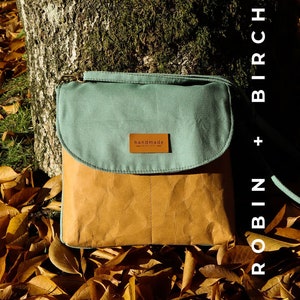 May include: A teal and brown crossbody bag with a flap closure and a leather tag that says "handmade". The bag is sitting on a bed of fallen autumn leaves.