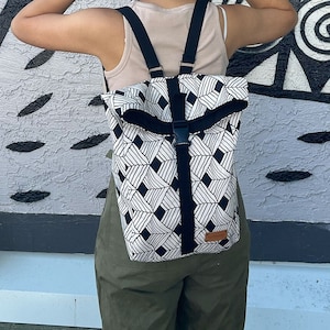 Op de afbeelding: Een zwart-wit geometrisch gepatroneerd rugzak met zwarte banden en een bruin leren patch. De rugzak wordt gedragen door een persoon met lang bruin haar.